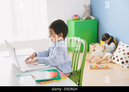 Elementary age boy doing his homework at his desk with his sister reading on the floor in the background Stock Photo