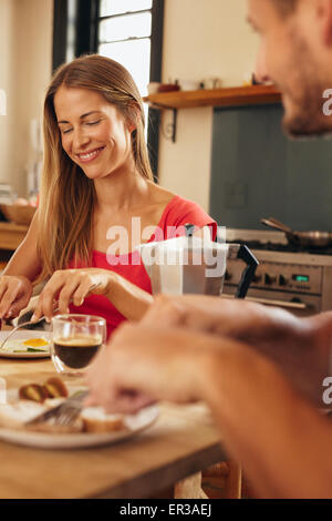Happy young couple having breakfast together in kitchen. Focus on woman smiling while eating and man in front is out of focus si Stock Photo