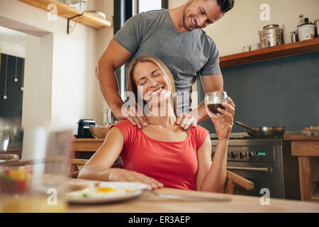 Happy young woman sitting at breakfast tablet holding cup of coffee getting a shoulder massage from her boyfriend. Young couple Stock Photo