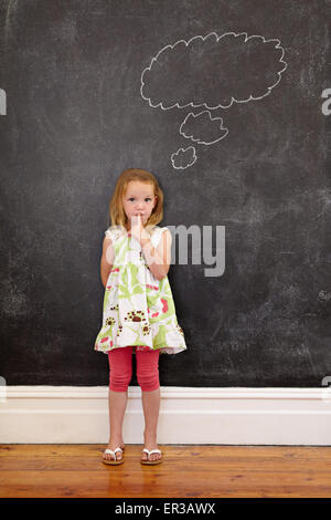 Cute little girl with her finger on lips and a thought bubble on blackboard. Little girl doing a please keep quiet gesture towar Stock Photo