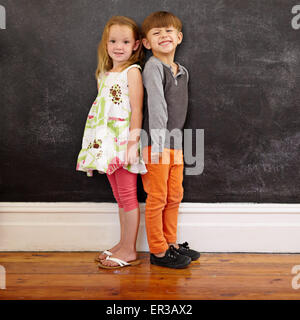 Little boy and girl standing back to back in front of blackboard looking at camera smiling. Full length image of two innocent li Stock Photo