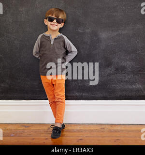 Full length shot of stylish little boy standing in front of the blackboard. Caucasian kid wearing sunglasses  posing at camera. Stock Photo