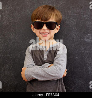 Photo of adorable young happy boy looking at camera with sunglasses. Little kid smiling with his arms crossed against blackboard
