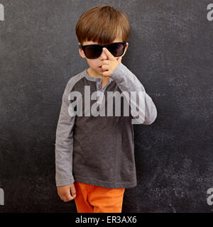 Portrait of little boy wearing sunglasses looking at camera. Small kid in front of a blackboard adjusting his shades. Stock Photo
