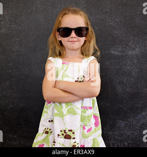 Portrait of cute little girl wearing sunglasses against a black wall. Young girl standing in shades with her hands folded. Stock Photo