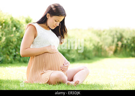 Pregnant Woman Sitting On Grass Outdoors Holding Bump Stock Photo