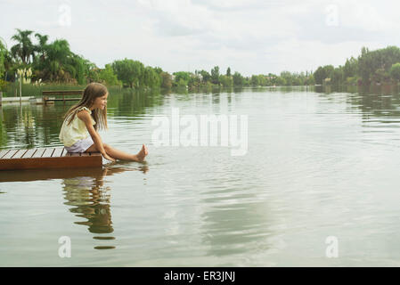 Girl sitting on dock with feet dangling in lake, portrait Stock Photo