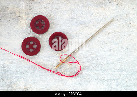 Sewing needle threaded with a red thread ready for sewing. Next , three buttons. Scene on a white wooden table . Top view. Stock Photo