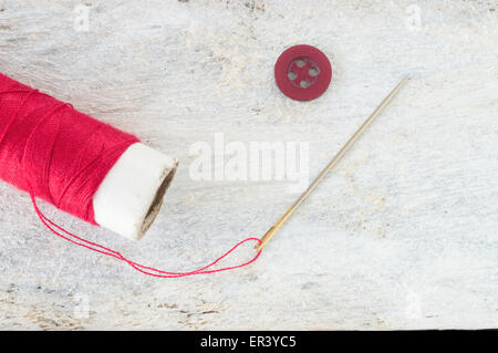 Sewing needle threaded with a red thread ready for sewing. Next , a button. Scene on a white wooden table . Top view. Stock Photo