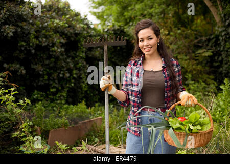 young woman in the garden holding a basket of fresh vegetables and a rake