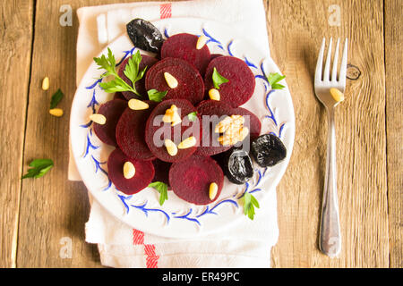 Boiled sliced beetroots (salad) on plate with pine nuts, walnuts, prunes (plumes) and parsley over rustic wooden table. Stock Photo