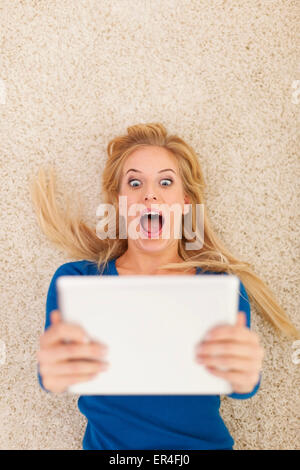 Surprise young woman lying on carpet and using digital tablet Stock Photo