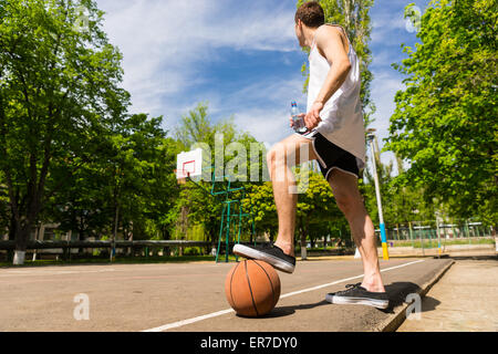 Low Angle View of Young Athletic Man Standing on Side Lines of Basketball Court with Foot on Top of Ball Preventing it from Roll Stock Photo