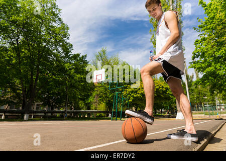 Low Angle View of Young Athletic Man Standing on Side Lines of Basketball Court with Foot on Top of Ball Preventing it from Roll Stock Photo
