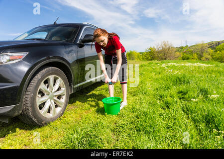 Woman with Green Bucket Wringing Out Soapy Sponge and Washing Black Luxury Vehicle in Green Field on Bright Sunny Day with Blue Stock Photo