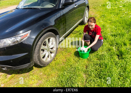 High Angle View of Woman with Green Bucket Wringing Out Soapy Sponge and Washing Black Luxury Vehicle in Green Field on Bright Sunny Day with Blue Sky. Stock Photo
