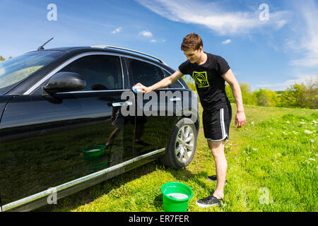 Full Length of Man Washing Car with Soapy Sponge in Green Grassy Field on Bright Sunny Day with Blue Sky Stock Photo