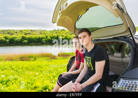 Couple Sitting on Rear Tailgate Bumper of SUV Vehicle Parked in Green Field by River on Sunny Day and Looking at Camera Stock Photo