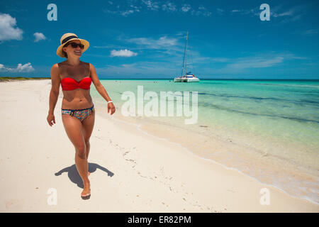 A young woman wearing a bikini and sun hat walks a sandy beach. Stock Photo