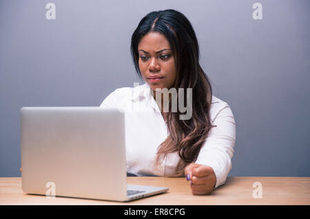 Angry african businesswoman sitting at the table and using laptop over gray background Stock Photo