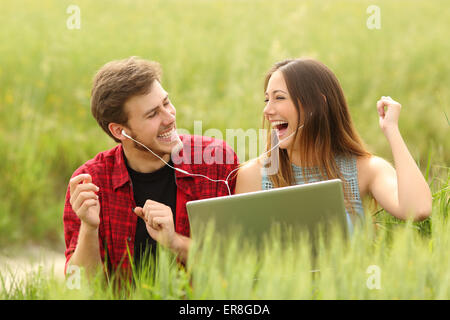 Funny couple singing and listening music from a laptop in a green field Stock Photo