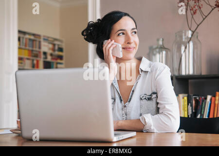 Beautiful young woman using mobile phone with laptop on table at home Stock Photo