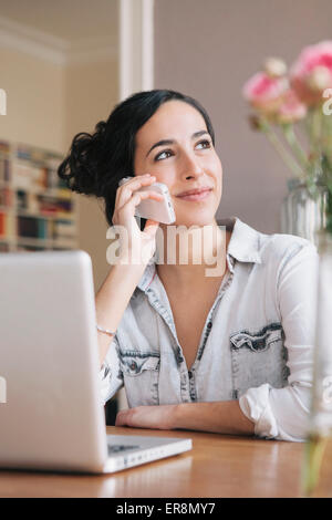 Beautiful young woman using mobile phone with laptop on table at home Stock Photo
