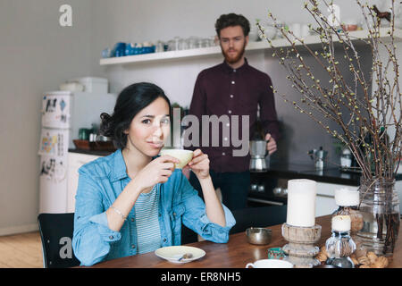 Portrait of young woman having coffee at kitchen table with man in background Stock Photo