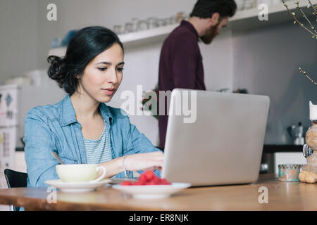 Young woman using laptop with man in background at kitchen