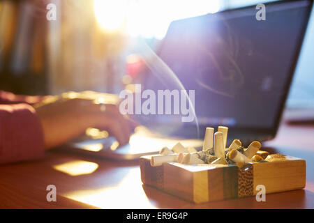 Close up of ashtray full of cigarette, with man in background working on laptop computer and smoking indoors on early morning. Stock Photo