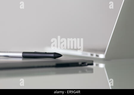 Still life of digital graphic tablet and stylus pen on desk, with laptop computer in background. Reflections on table and copy s Stock Photo