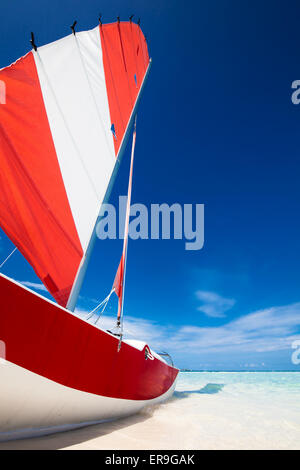 Sailing boat with red sail on a beach of deserted tropical island with shallow blue water
