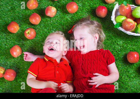Child eating apple. Little girl and baby boy play peek a boo holding fresh ripe apples. Kids eat snack relaxing on a lawn. Stock Photo