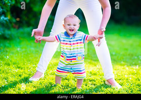 Baby boy making his first steps. Mother holding her child. Kid walking on a lawn in a sunny summer garden. Kids learning to walk Stock Photo