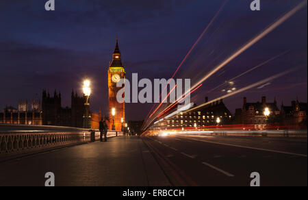 A couple kiss on the bridge as a double-decker bus zooms past the clock tower of the Palace of Westminster (Big Ben) in London. Stock Photo