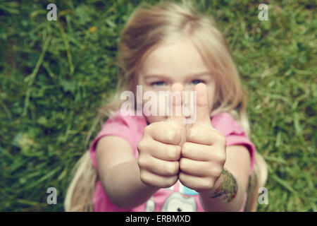 child blond girl lying on green grass in summer park showing thumbs up gesture using both hand Stock Photo