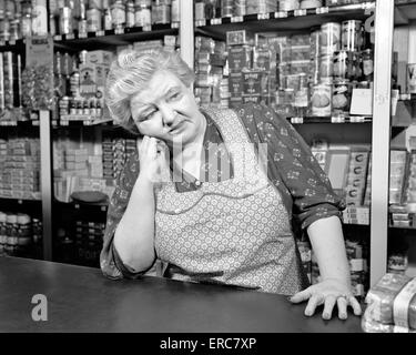 1930s 1940s SAD SENIOR WOMAN CLERK GROCERY STORE WEARING APRON CANNED GOODS ON SHELVES LEANING COUNTER Stock Photo