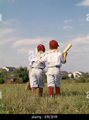 1960s TWO BOYS BROTHERS TEAMMATES WEARING LITTLE LEAGUE BASEBALL UNIFORMS WALKING ARM IN ARM AWAY FROM CAMERA Stock Photo