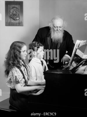 1930s 1940s BOY GIRL SEATED AT PIANO WITH ELDERLY MAN TEACHER WATCHING THEM PLAY Stock Photo