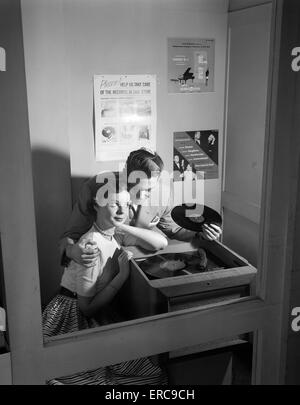 1950s COUPLE SITTING IN STORE RECORD LISTENING BOOTH MAN WITH ARM AROUND WOMAN HOLDING 78 RPM Stock Photo