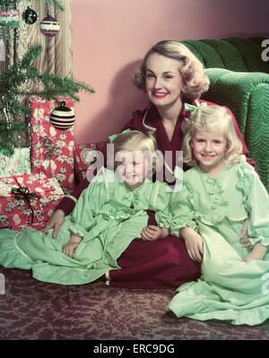 1950s SMILING MOTHER AND TWO DAUGHTERS IN NIGHTGOWNS POSING SITTING ON FLOOR BY CHRISTMAS TREE LOOKING AT CAMERA Stock Photo