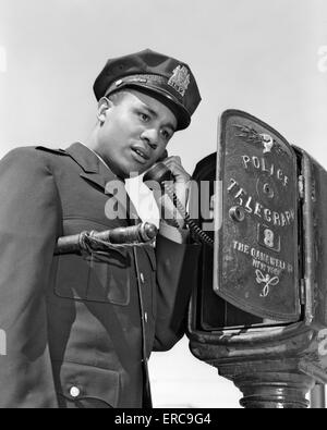 1960s AFRICAN AMERICAN POLICE OFFICER MAKING CALL AT POLICE BOX TELEPHONE NIGHT STICK TUCKED UNDER HIS ARM Stock Photo