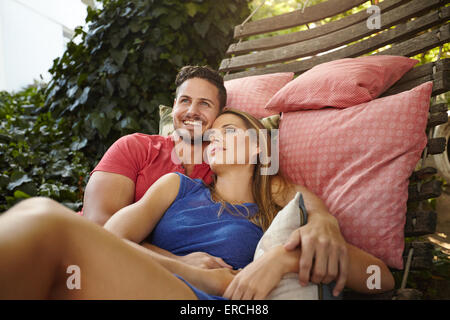 Portrait of happy and peaceful young couple relaxing on hammock in backyard. Man smiling with woman looking away. Stock Photo