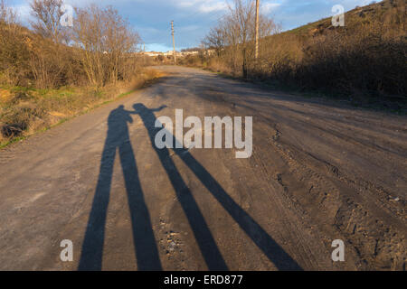 Tall Shadows of Two People or Couple Stretching Along Dirt Road in Rural Area at Sunrise or Sunset
