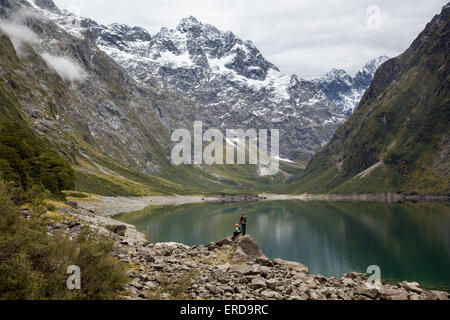 Two walkers look out over Lake Marian in the Darran Mountains of Fjordland in South Island New Zealand Stock Photo