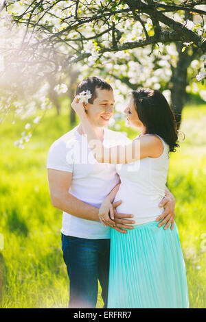 Portrait of happy pregnant family. Pregnant woman and man standing in blooming spring garden in sunny day. Two people in love. Stock Photo
