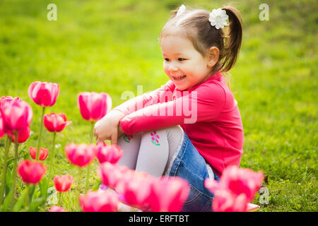 cheerful little girl sitting in grass looking at tulips . kid plays outside Stock Photo