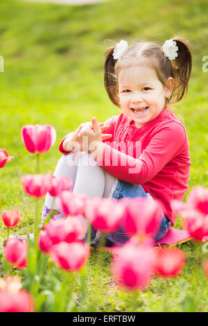 cheerful little girl sitting in grass looking at tulips . kid plays outside Stock Photo