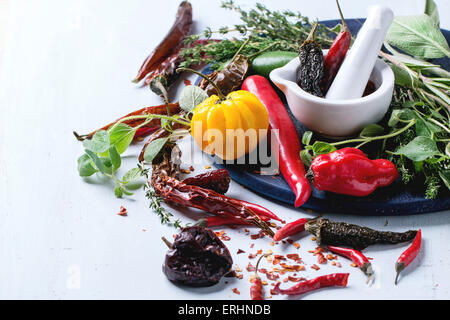 Assortment of fresh, dryed and flakes hot chili peppers and fresh herbs with white ceramic mortar on dark blue cutting board ove Stock Photo