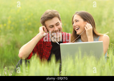 Happy couple or friends sharing music from a laptop sitting in a green field and looking each other Stock Photo